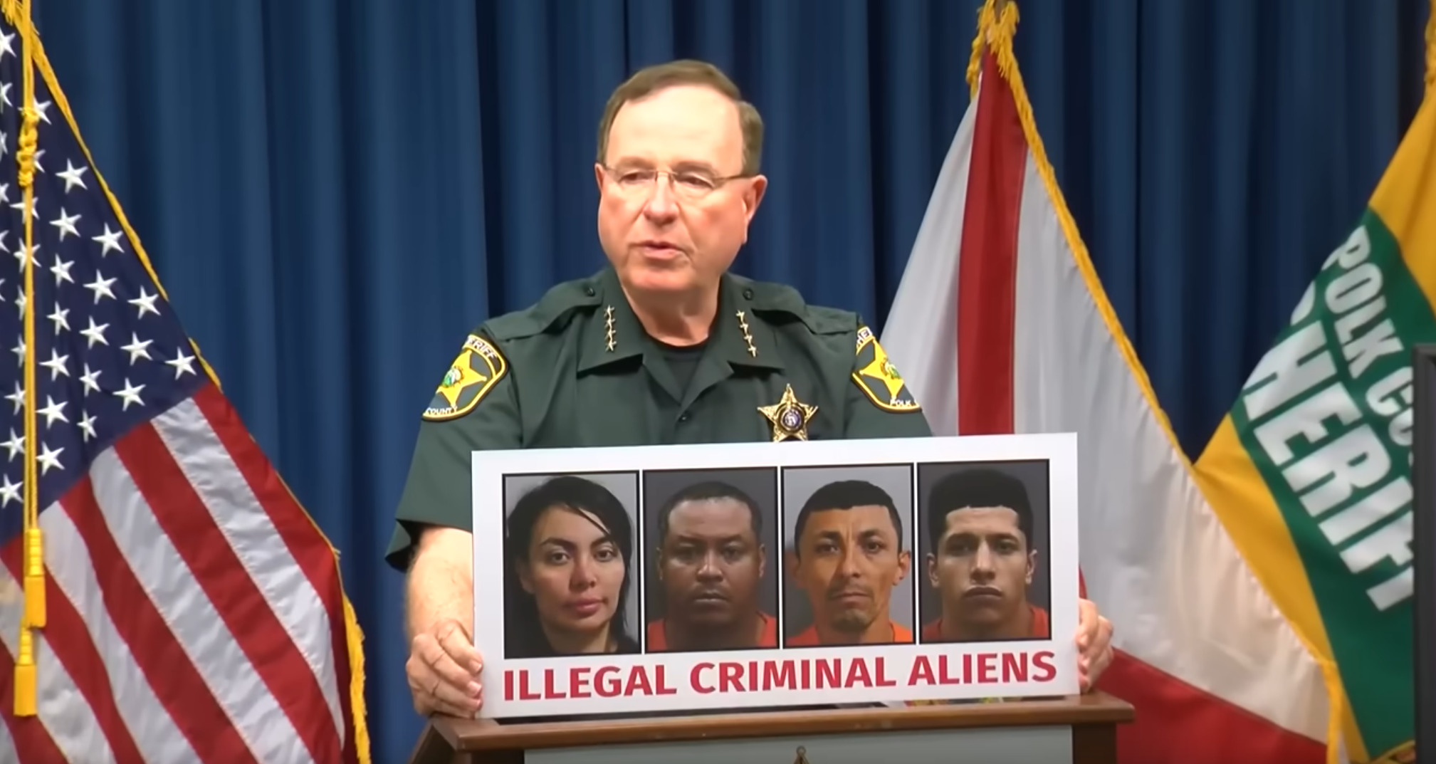 a US police person at a press conference holding a placard with four mugshots and the heading "ILLEGAL CRIMINAL ALIENS"